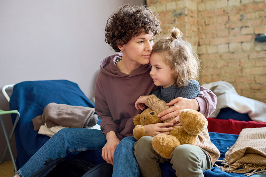 Young woman giving hug to her cute little son with brown soft teddy bear while both sitting on sleeping place prepared