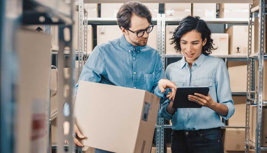 Female Inventory Manager Shows Digital Tablet Information to a Worker Holding Cardboard Box, They Talk and Do Work. In the Background Stock of Parcels with Products Ready for Shipment.